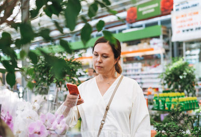 Middle aged woman in white dress take photo on mobile of green potted house plants at garden store