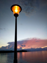 Illuminated street light by lake against sky at sunset