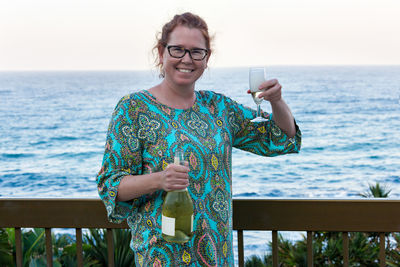 Portrait of smiling mature woman holding champagne flute against sea