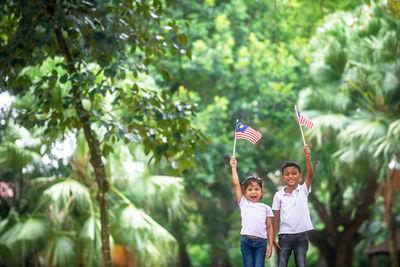 Portrait of siblings holding malaysian flag while standing against trees at park