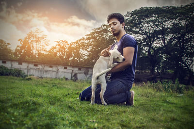 Side view of man stroking dog while kneeling on field against sky