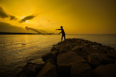 Silhouette fisherman throwing fishnet in sea against sky during sunset