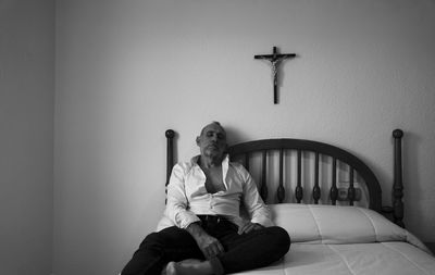 Monochrome of adult man in white shirt in room lying on bed with religion cross on wall