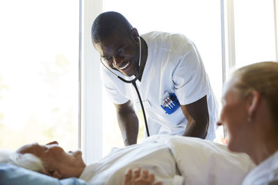 Happy nurses talking to senior man lying on bed in hospital ward