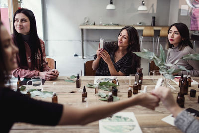 Smiling young woman photographing through smart phone while sitting at table amidst multi-ethnic colleagues