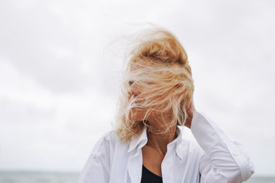 Portrait of elegant blonde woman in white shirt on sand beach at storm sea at windy weater