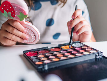 Crop unrecognizable kid with applicator and mirror making up face at table with assorted cosmetic products in house