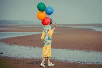 Rear view full length of young woman holding colorful balloons at beach