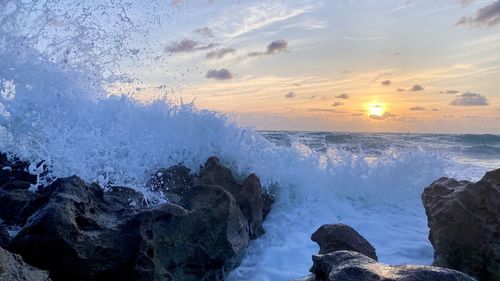 Waves splashing on rocks at shore against sky during sunset