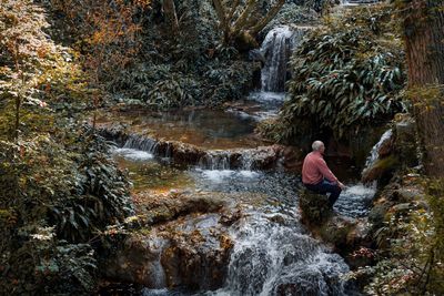 Side view of man sitting on rock by waterfall in forest