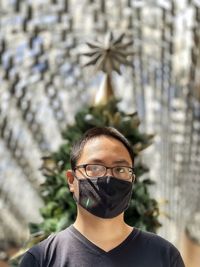 Portrait of young man wearing eyeglasses and face mask against ceiling and christmas tree.