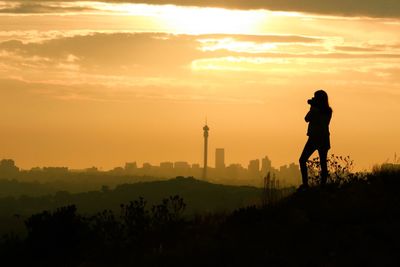 Silhouette woman photographing while standing on grass against sky during sunset