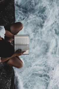 High angle view of man reading book on rock formation by sea