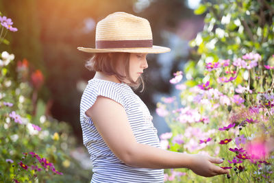 Side view of woman wearing hat standing against pink flowering plants