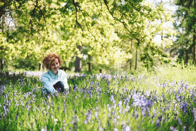 Thoughtful smiling woman sitting amidst plants in forest on sunny day