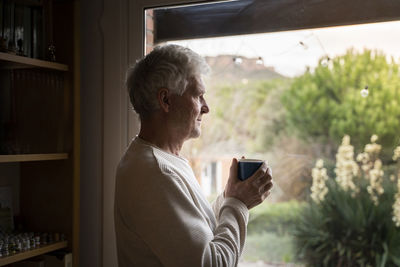 Senior man with coffee cup looking through window while standing at home