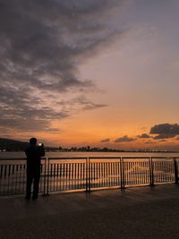 Rear view of man standing by railing against sky during sunset