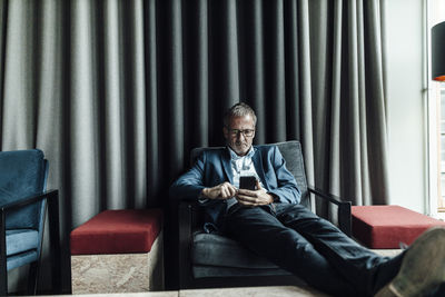 Serious businessman using mobile phone while resting on armchair in cafeteria at office