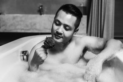 Shirtless man holding rose while siting in bathtub