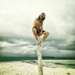 Man wearing a virtual reality headset while sitting on wood at beach against cloudy sky