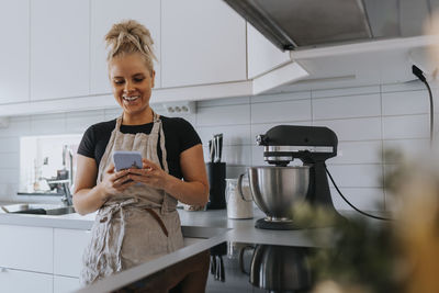 Smiling young woman in kitchen using cell phone