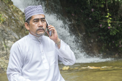 Thoughtful man talking on phone while sitting against waterfall