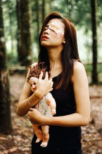 Young woman with wounded eye holding doll while standing in forest