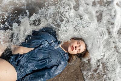 From above of calm female in wet shirt lying with closed eyes on sandy beach near waving sea on sunny summer day