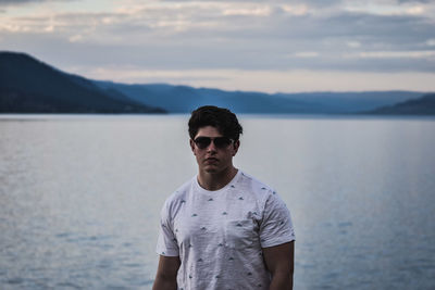 Portrait of confident young man wearing sunglasses against lake during sunset