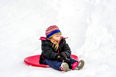 A little girl on a red saucer sled, laughs as her hat falls over her eyes 