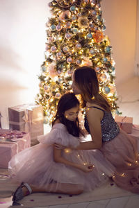 Baby girl with big eyes and a mom in a pink dress is sitting at the christmas tree