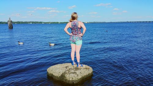 Rear view of woman with hands on hip standing on rock at sea