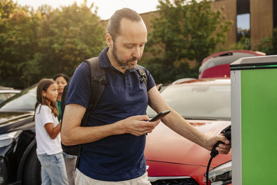 Man using smart phone while holding plug at car charging station