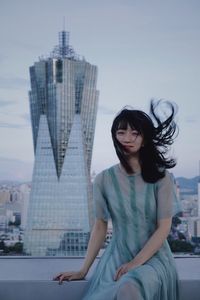 Young woman sitting against modern buildings in city against sky
