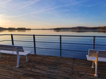 Empty chairs and table by lake against sky during sunset