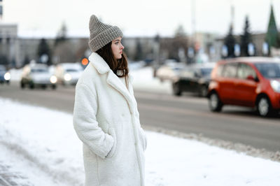 Girl in a fur coat walks along the road traffic of cars. the girl has a knitted hat on her head