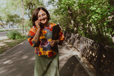 Cheerful young woman with glasses talking on a mobile phone in lush green park.