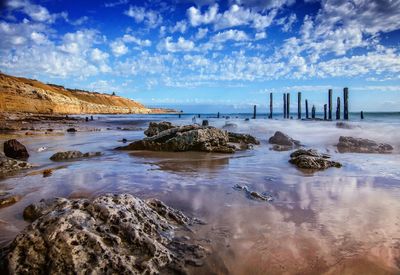 Rocks and wooden posts in sea against cloudy blue sky