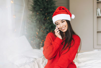 Young asian woman in red sweater and santa hat using mobile in room with christmas tree at home