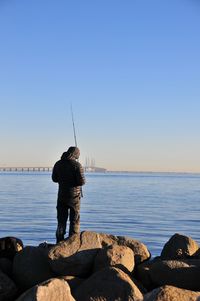 Rear view of man fishing at sea shore against clear sky