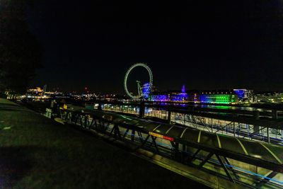 Illuminated ferris wheel by river against sky in city at night