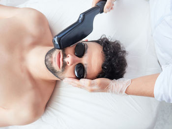 From above cropped faceless master using laser apparatus on male face removing beard face during procedure in salon