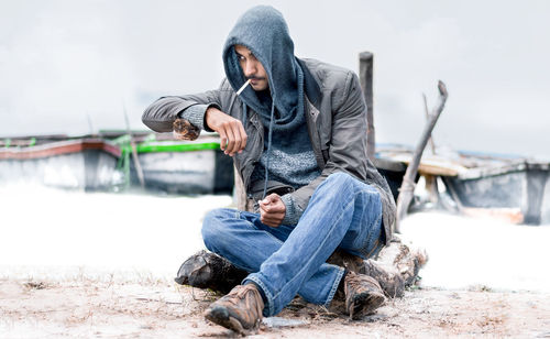 A man sitting on wood and looking right side with cigarette putting on hoddy.