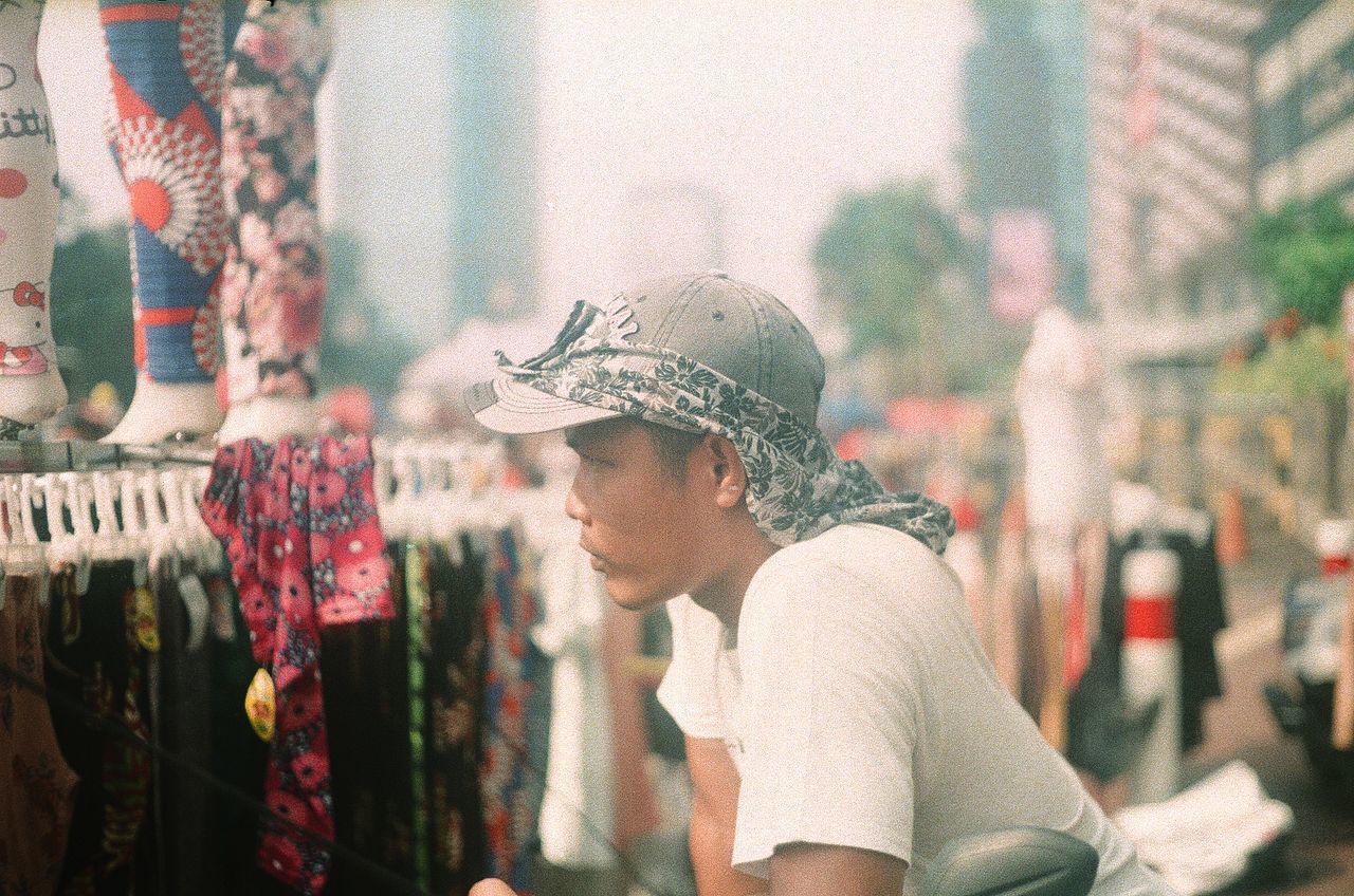 one person, hat, side view, real people, focus on foreground, clothing, day, lifestyles, incidental people, looking, leisure activity, adult, headshot, casual clothing, waist up, retail, men, young adult, looking away, outdoors, retail display, profile view, contemplation