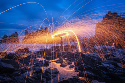 Man spinning wire wool at rocky beach against cloudy sky during night