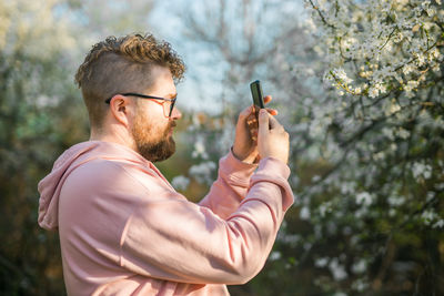 Side view of young man photographing while standing against trees