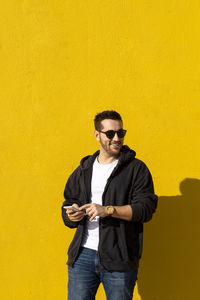 Bearded man with sunglasses standing against yellow wall, using phone