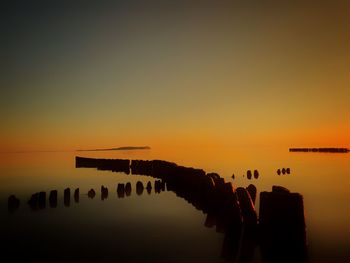 Silhouette wooden posts in sea against orange sky