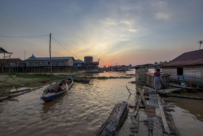Boats moored on river by buildings against sky during sunset