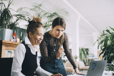 Businesswoman looking while smiling female colleague using laptop in creative office